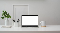 Cropped shot of modern workspace with blank screen laptop, frame, pencils, coffee cup and vase on white table with white wall background    