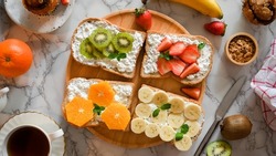 Yummy healthy fruits on a toast with cottage cheese on wooden board, a cup of coffee, banana, orange, kiwi, strawberry, orange and granola bowl on white marble background. top view