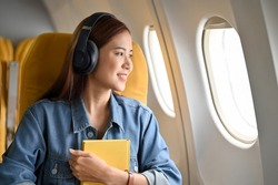 Happy and cheerful young asian woman with headphones looking the view outside of the plane window. Female passenger image