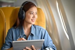 Happy and cheerful young asian woman with headphones using a portable tablet and looking the view outside of the plane window. Female passenger image
