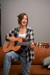 A talented and attractive young Asian woman sings while playing her acoustic guitar in the living room.