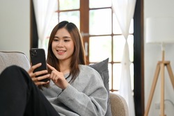 Attractive and happy young Asian female using her smartphone to scroll through social media while relaxing on her comfortable sofa in her living room. 
