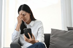 Stressed young Asian female using her smartphone while sitting on sofa, suffering from headache or eye strain, feeling upset or dissatisfied with something.