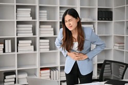 Sick and unwell millennial Asian businesswoman suffering from stomachache or abdominal pain in her office.