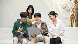Group of happy young Asian college students sitting on a bench, looking at a laptop screen, discussing and brainstorming on their school project together.