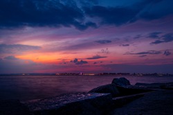 Watching the sunset on Breezy Point Beach on the end of one summer warm day. Dramatic cloudy dark blue sky and orange purple touch of the sun in the background. Big rocks in the foreground.