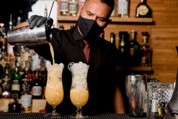 Young professional bartender is preparing cocktail for his clients at work with medical mask and protection against coronavirus COVID-19. profession concept
