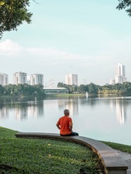 A senior citizen in orange shirt meditating during the early morning in a park while sitting facing the lake
