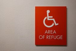 A sign in a hallway in an apartment complex that says Area of Refuge with a wheelchair symbol on it that is an emergency gathering area for disabled people.