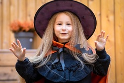 Happy scary Halloween kid! Cute little witch girl with pumpkin, bat say boo. Beautiful young child in orange, black costume with hat frighten outdoors.