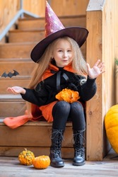 Happy scary Halloween kid! Cute little girl witch with candy bucket, pumpkins, bat say boo. Beautiful young child in orange, black costume with hat sitting on ladder home frighten outdoors.