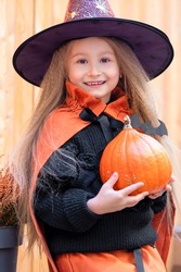 Happy scary Halloween kid! Cute little girl witch with candy bucket, pumpkins, bat say boo. Beautiful young child in orange, black costume with hat sitting on ladder home frighten outdoors