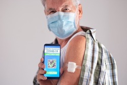 Portrait studio shot of old caucasian senior man patient wearing face mask showing green pass app after booster third doses of covid-19 coronavirus vaccine, looking at camera