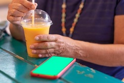 Woman holds in her hands an orange healthy smoothie with orange, papaya and maracuja peach. Mature elderly lady relaxing sitting at wooden table looking at smart phone with green screen