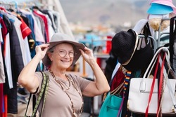 Attractive elderly woman at the flea market looking for second hand  clothes, caps, shoes, bags. Zero waste shopping, eco friendly concept, sustainable lifestyle.