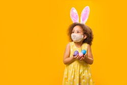 A cheerful girl with rabbit ears on her head and a protective mask with a basket of colored eggs in her hands on a yellow background. She looks away. Funny happy baby. Covid Easter to the child.