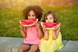 Funny Black and European girls are eating watermelon outdoors in the hot summer. Smiling children in light dresses enjoy healthy food