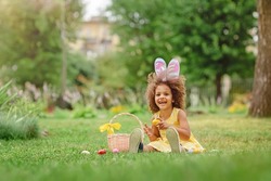 Little Black girl wear bunny ears and gathering Easter eggs on Easter egg hunt in garden