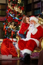 Santa Claus tells a funny story to a cute little girl on the holiday of Christmas. A smiling little girl hugging Santa Claus against the background of the Christmas Tree lights.