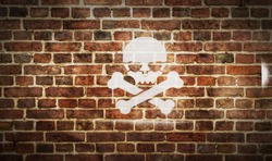 Skull piracy symbol spray painted on the brick wall. Graffiti art concept of piracy, danger and warning. Urban abstract artwork. Airbrush paint with sign template in hand.