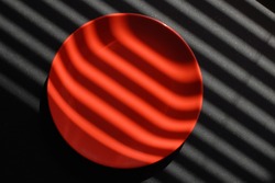 A round flat coral-red ceramic plate on a black plastic background is illuminated by sunlight through the blinds. Hard light draws clear stripes and creates an abstraction.