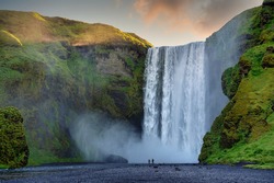 Two male and female tourists are walking at Skogafoss Falls in southern Iceland. In the morning, the sun rose from behind the mountains with green grass. This is a popular tourist destination.