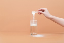 Woman pours collagen powder or protein in a glass of water on a beige background. A healthy and anti aging supplement. Copy space
