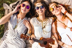 Three cute hippie girl lying on the plaid outdoors, best friends having fun and laughing, play ukulele, sunglasses, feathers in their hair, bracelets, flash tattoo, indie, Bohemia, boho style top view