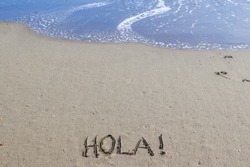 The word hello in Spanish with an exclamation point (Hola!) on a golden sandy beach with blue ocean waves rolling in from above.  Peaceful, tranquil, tropical, vacation concepts.