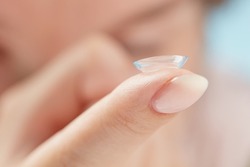 Soft contact lens on female finger against the blurred face, close up. Medicine and vision concept. Woman putting soft contact lens into eye. Shallow depth of field.