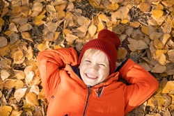 Child in an orange jacket lies on the fallen leaves in the autumn forest. Rest in the fall in nature.