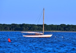 A wooden Sail boat and red buoy with sails down and anchored in a beautiful blue bay.The bay is Lewis Bay in Cape Cod Massachusetts.