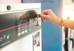 Female hand ready to insert a token into the slot of vintage chrome coin on the washing machine. Asian woman hand putting coin in washer machine for operated, inside laundrette, closeup