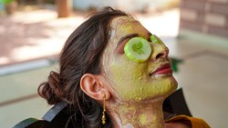 A beautiful woman with facial clay mask on face and fresh cucumber on eyes relaxing at Home. Portrait of beautiful spa girl. Beauty, healthcare. Multani Mitti and green Face Pack in India