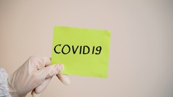 Close up of hand in disposable protective gloves picks up a sheet of bright paper with the inscription Covid 19. Coronavirus epidemic. Pandemic of airborne viral disease