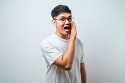 Young handsome asian man wearing casual shirt and glasses over white background hand on mouth telling secret rumor, whispering malicious talk conversation