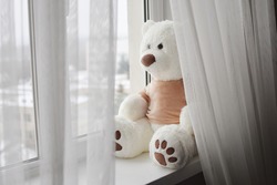 Big white teddy bear toy is sitting at the window, looking out. Selective focus. 