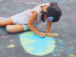 Little girl with pigtails sits and uses colorful chalks draws and paint green earth on the road floor. Earth day concept. Kids outdoor activities concept.