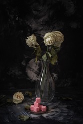 Squared pieces of rose colored sweets lying on small steel plate, three faded roses in glass vase and one rose on the table, dark background