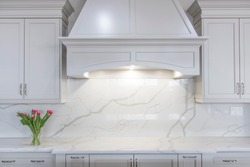 A modern white kitchen with a traditional touch. White marble-looking quartz countertop and backsplash accompanied by white cabinetry and custom made range hood.