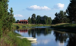 A picture of rural Swedish countryside. A traditional red house (stuga) in the background, and a boat on a lake. An idyllic landscape of Dalarna, Sweden. A summer view of Malung.