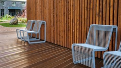 Resting point inside residential area courtyard in rainy day.Two wet metal chairs with raindrops of modern design close-up on a background of wet wooden shield.Comfortable urban public furniture.