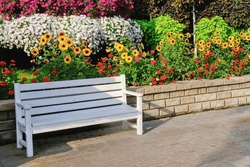 White wooden bench beside path in garden with blossom flowers and trees in the background.Bench in public garden. Urban street furniture.