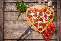 Valentines day heart shaped pizza with pepperoni, cherry tomatoes, mozzarella and parsley on vintage wooden table background. Symbol of love.