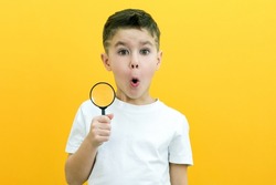 Positive curious schoolboy in white t-shirt looking at camera through magnifying glass on yellow background