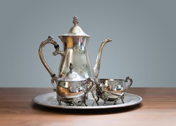 Ornate 4-piece tea set on table. Silver or silver plated tea pot, sugar bowl and cream or milk jug. Ornamental silverware to serve hot tea or coffee. Selective focus.