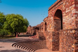 Huge gate structure at a fort made with red sandstone called Talaqi Darwaza near an amphitheatre with blue sky and green trees. A famous monument known as Purana Qila or Quila in Delhi, India.