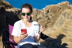 Beautiful girl traveler using smartphone, holding iron mug cup of coffee, sitting in touristic chair on cliff near ocean outdoors.Checking social media, chatting with friend,reading news,sharing data