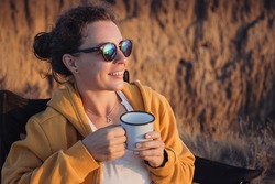 Happy young woman traveler in sunglasses holding iron mug cup with tea or coffee, enjoying ocean sunset scenery in nature landscape, sitting in camp. Travel camping and adventure lifestyle concept.