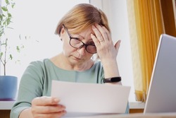 Shocked worried mature woman reading paper document or postal letter feeling frustrated by unexpected news sitting at home indoor. Received bank account balance or bad notice, fine, bankruptcy